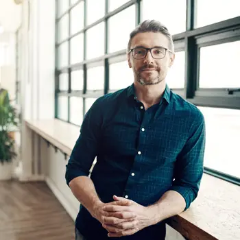 man with blue shirt and glasses staring into the camera while slightly smiling. he is leaning against the window sill.